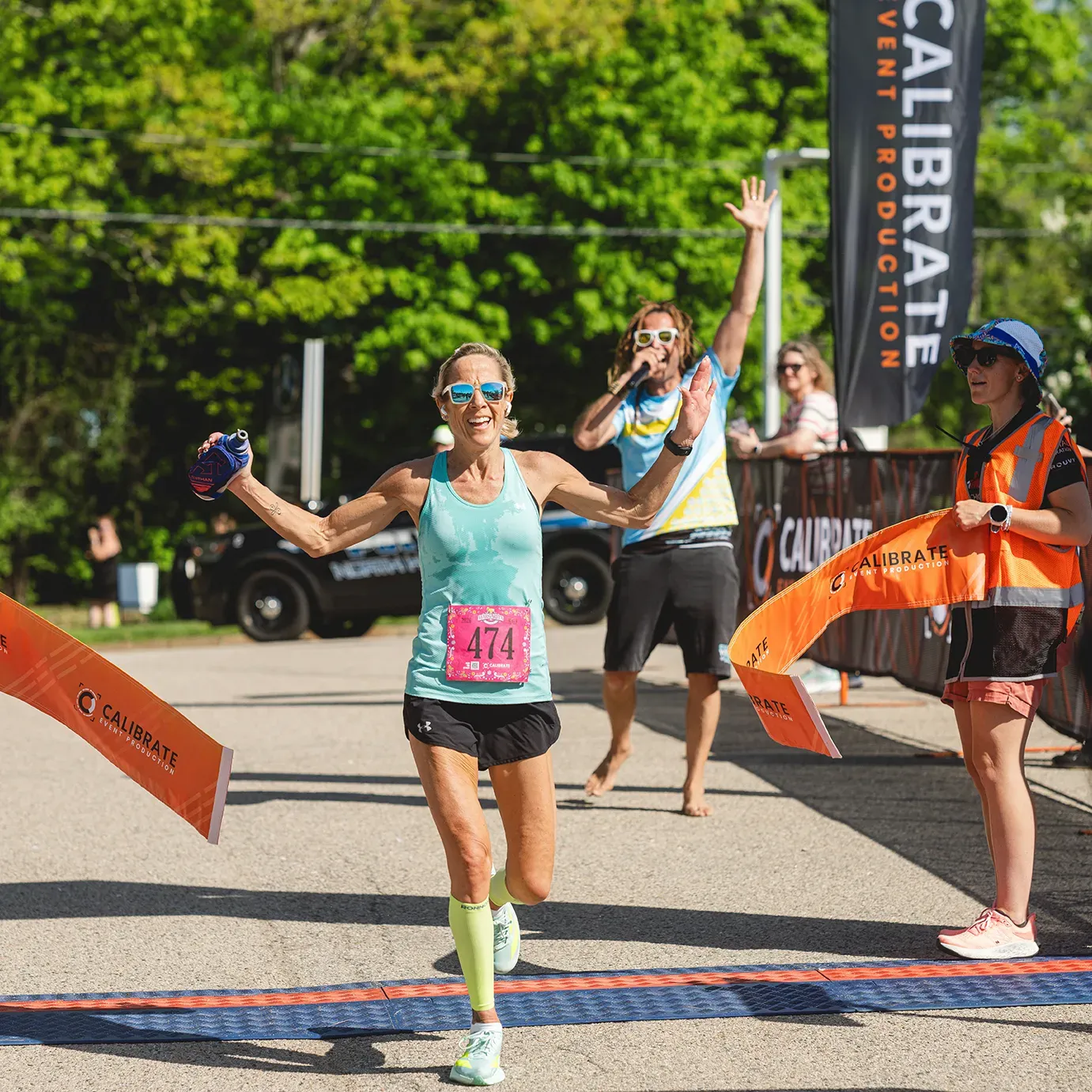 A female runner with arms outstretched crosses the finish line, smiling, with a race announcer and official in the background.