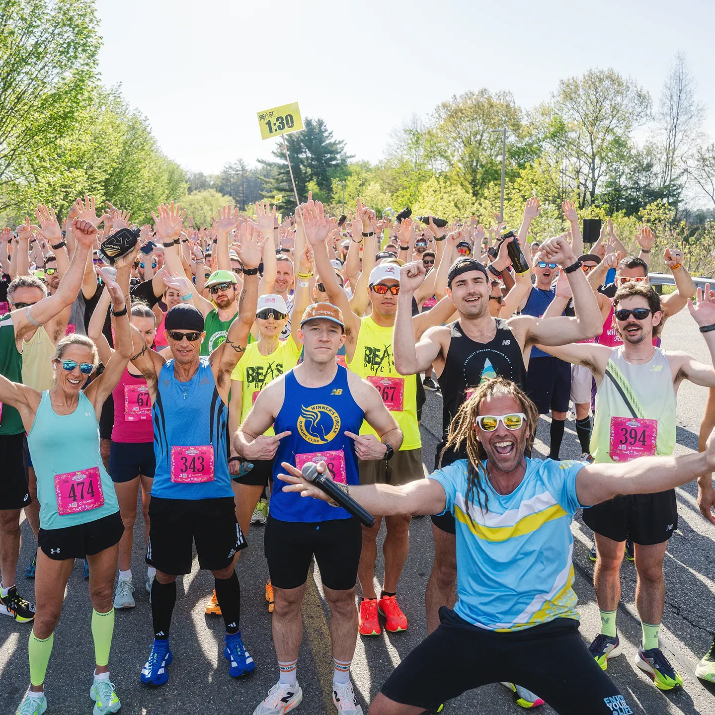 A large group of runners with their hands raised in excitement at the start line of a race, with one man in the foreground wearing sunglasses and a blue and yellow shirt, smiling broadly.