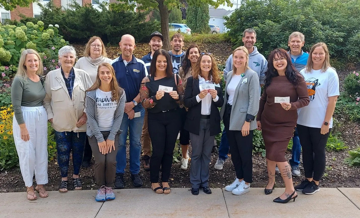 A group of people, some holding checks, pose for a photo outdoors with greenery in the background.