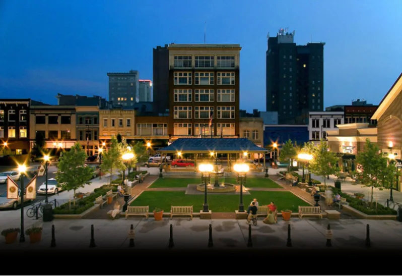 A picturesque view of a town square at dusk, with buildings, trees, and people relaxing on benches.