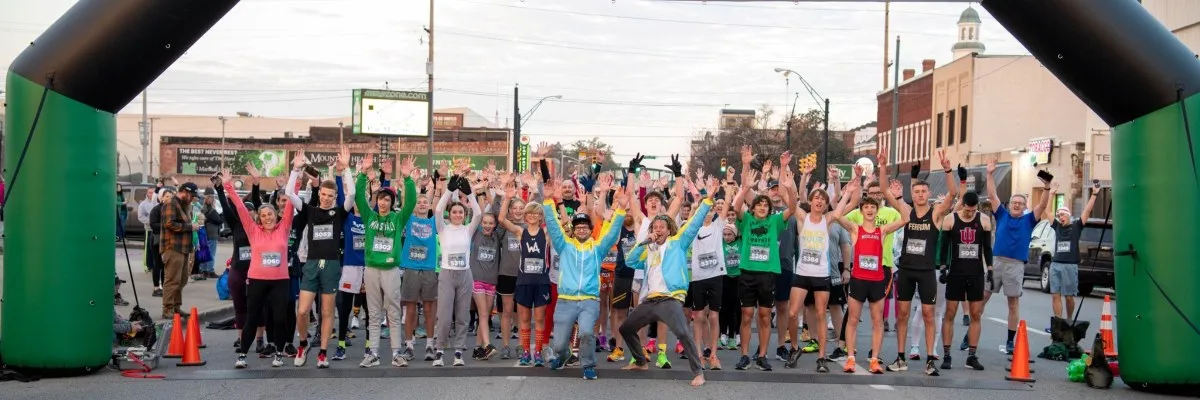 A large group of runners with their hands raised in excitement at the start line of a race, under a green and black inflatable arch.