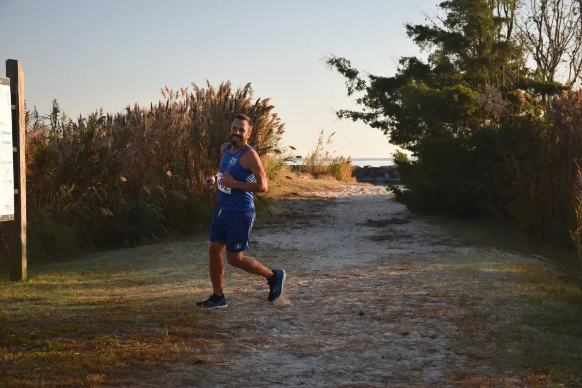 A male runner in a blue tank top and shorts runs along a sandy path next to tall grasses, with the ocean visible in the distance.