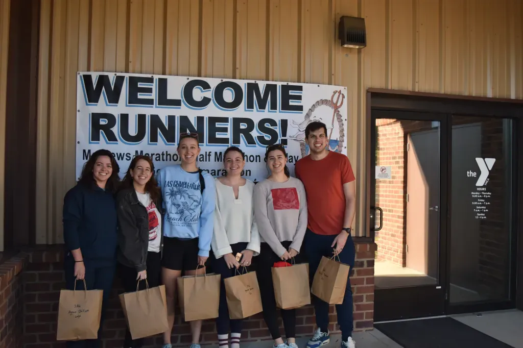 A group of six people, five women and one man, stand in front of a banner that says "WELCOME RUNNERS!" and hold brown paper bags.