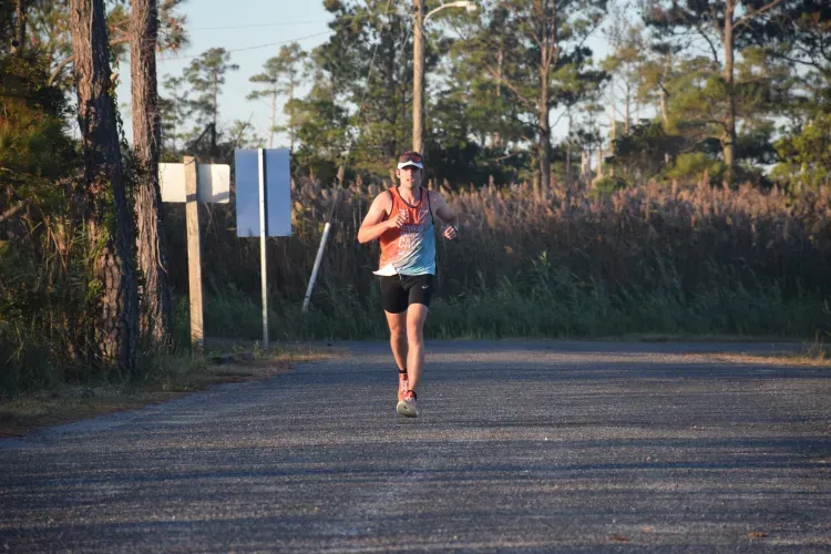 A male runner in a colorful tank top and black shorts runs down a paved road, with trees and tall grass on either side.