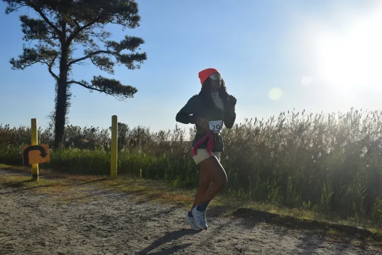 A runner wearing a bright orange beanie and sunglasses runs on a dirt path next to tall grass, with a tree and blue sky in the background.