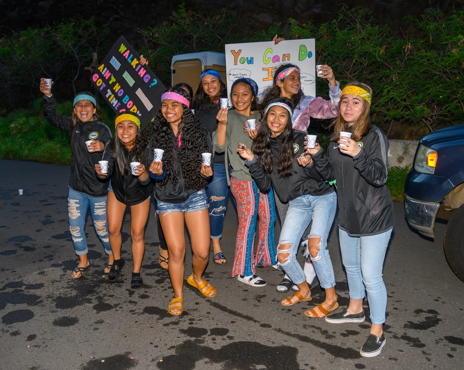 A group of young people, some holding cups and signs, pose for a photo. One sign reads 'WALKING? AIN'T NOBODY GOT TIME FOR THAT!' and another says 'You Can Do It!'