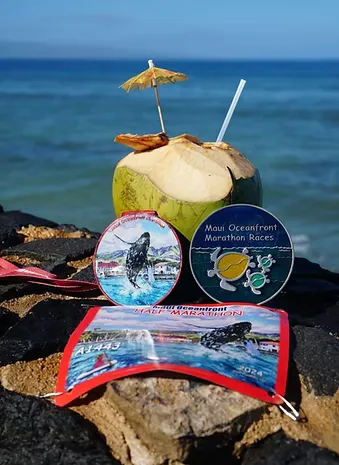 Maui Oceanfront Half Marathon medals and bib displayed on rocks with a coconut drink and the ocean in the background.