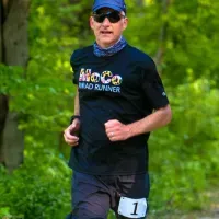 A male runner wearing a black t-shirt with colorful text and sunglasses, running on a wooded path.