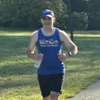 A male runner wearing a blue tank top and a blue cap runs on a paved path outdoors.