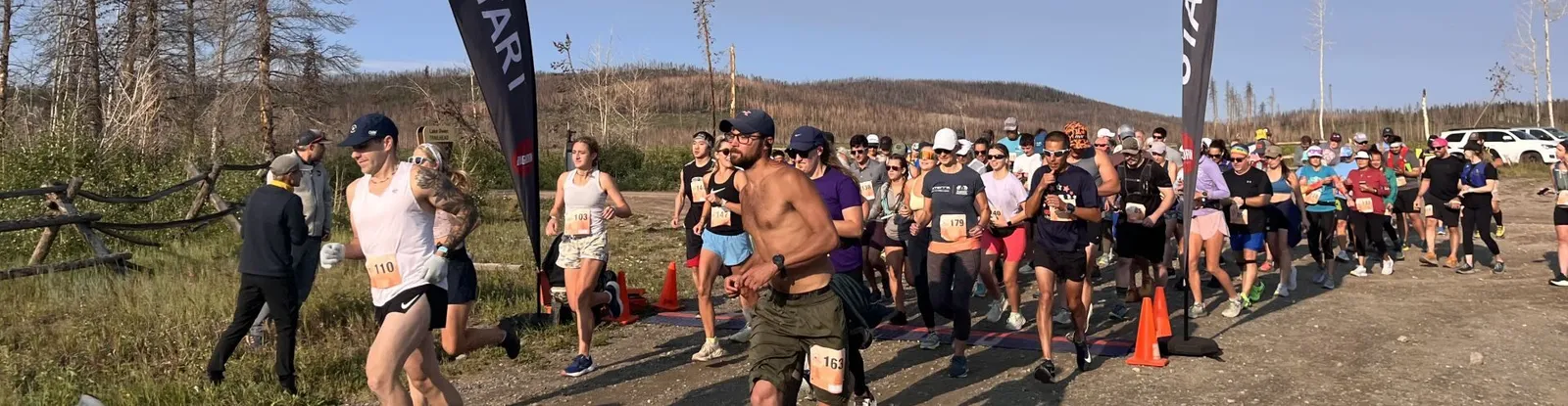A group of runners at the start line of a race, with trees and a banner in the background.
