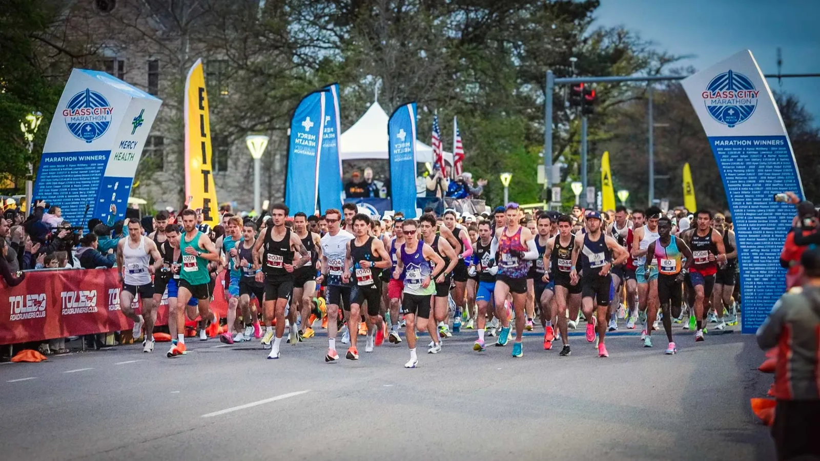 Runners surge forward at the start line of the Glass City Marathon, with spectators and race banners in the background.