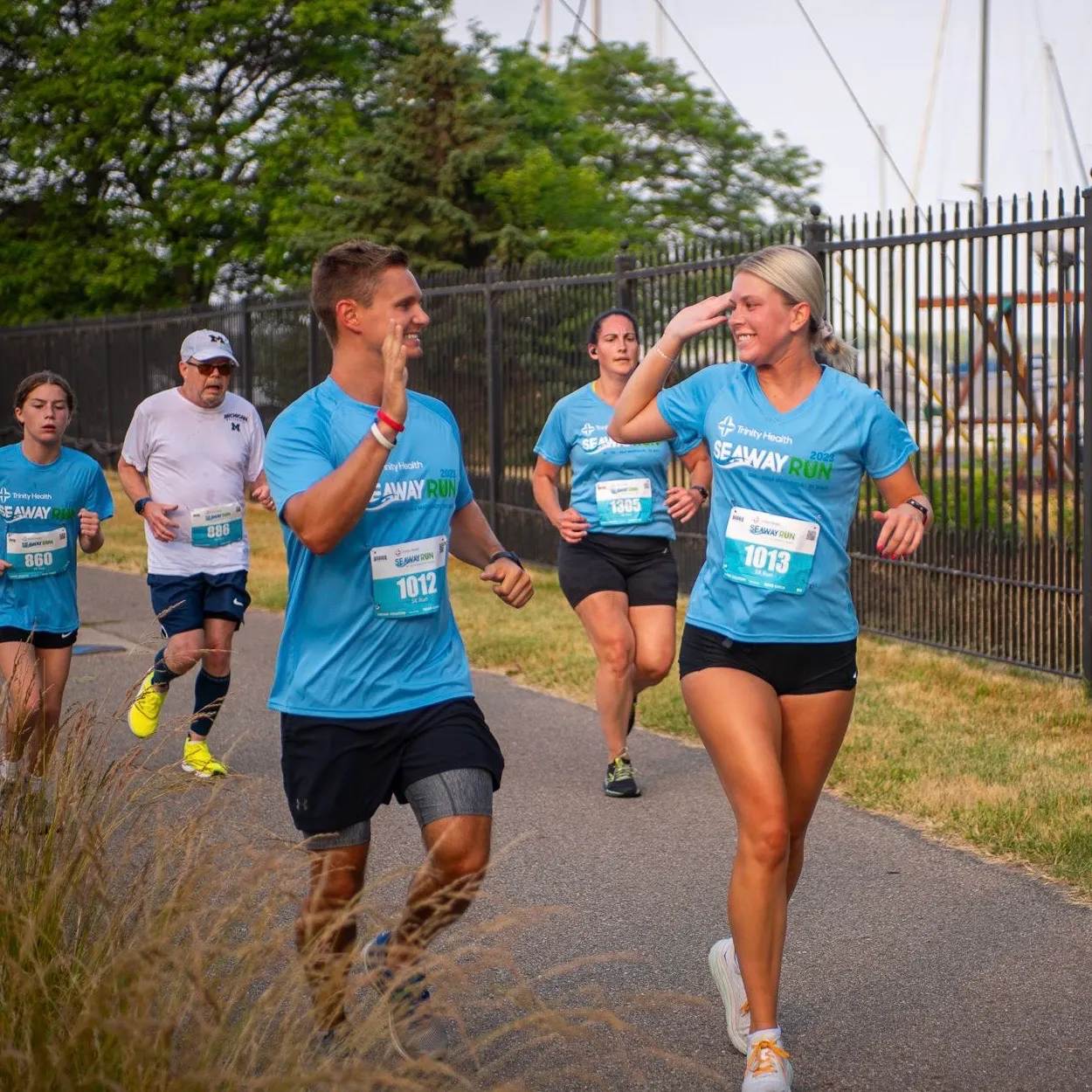 Runners in blue 'Seaway Run' t-shirts high-fiving and running on a paved path with a fence and trees in the background.