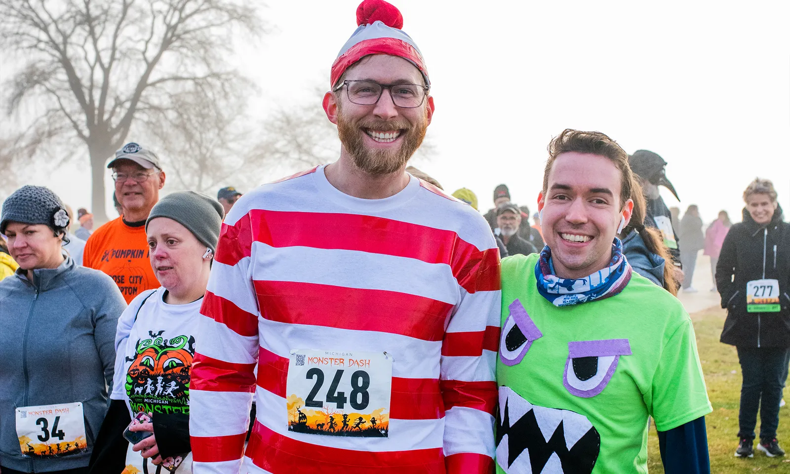 Two smiling participants in costumes at the Michigan Monster Dash race, one dressed as Waldo and the other in a green monster shirt, with other runners in the background.