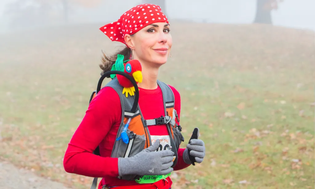 A female runner wearing a red long-sleeved shirt, a red polka-dot bandana, and a stuffed parrot on her backpack, gives a thumbs up while running in a foggy, outdoor setting.