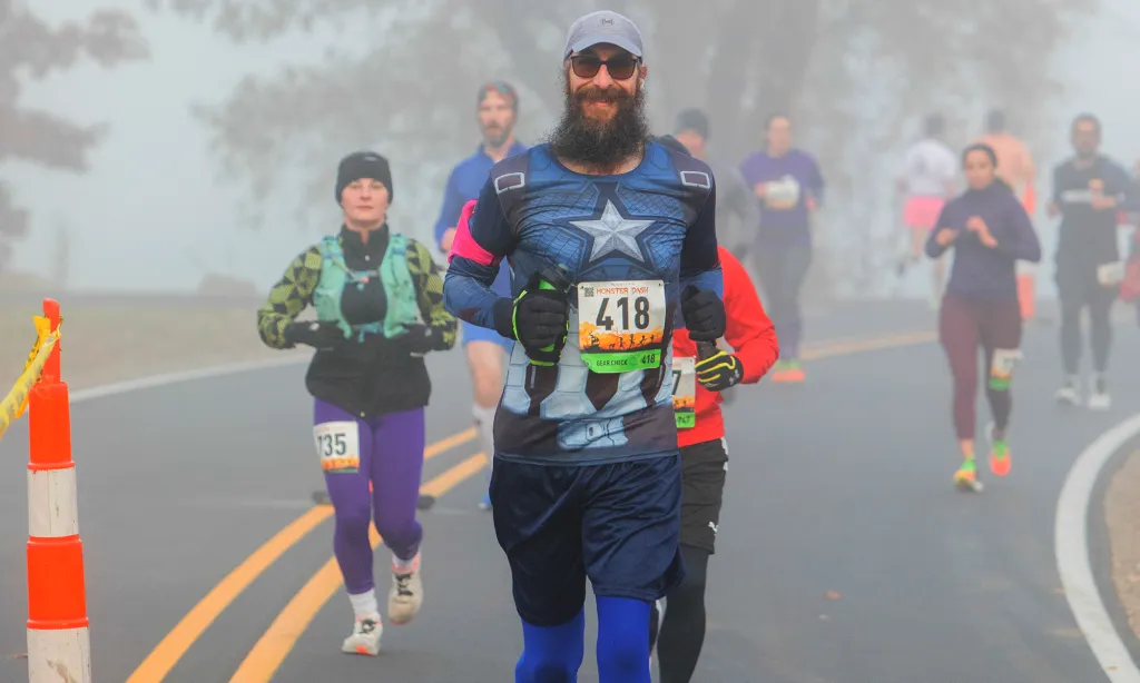 A runner dressed as Captain America smiles at the camera while participating in a foggy race, with other runners visible behind him.