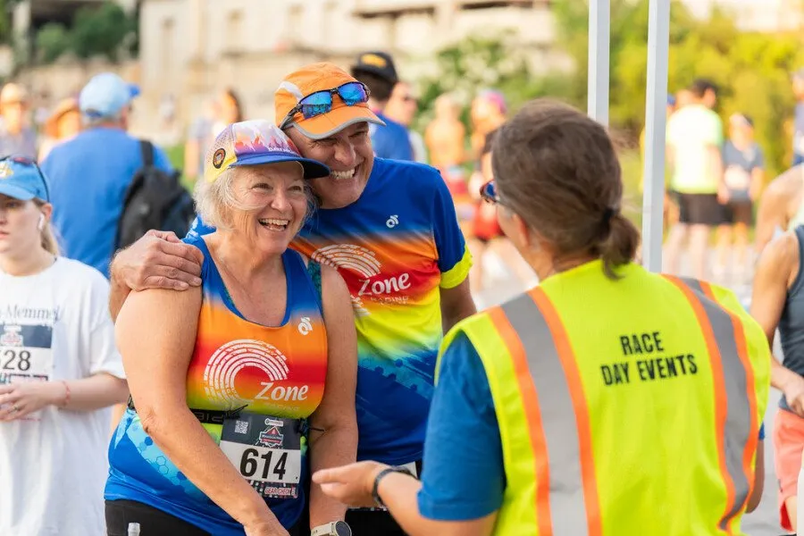 A smiling older couple, both wearing race bibs and athletic wear, are interacting with a race day volunteer wearing a bright yellow vest.