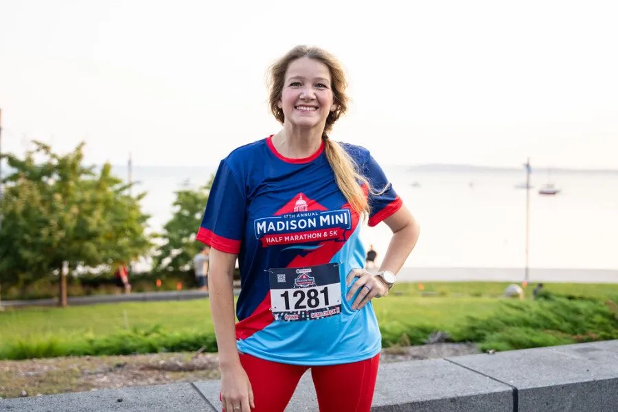 A smiling woman wearing a blue and red Madison Mini Half Marathon & 5K shirt and red leggings, with race bib number 1281, standing outdoors with a waterfront in the background.