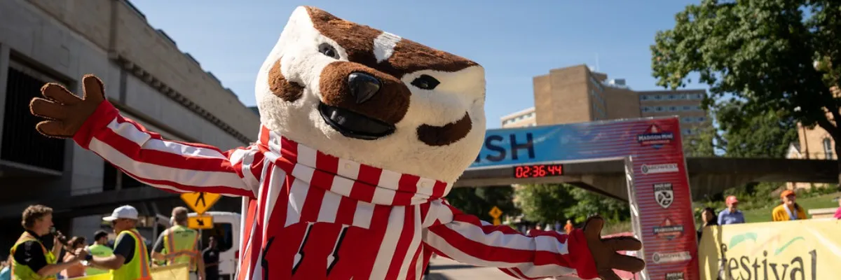 A mascot in a red and white striped uniform with outstretched arms stands in front of a finish line banner at a race.