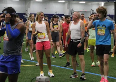 Runners in athletic wear gathered on a turf field, some wearing bib numbers, appearing to wait for a race to begin.