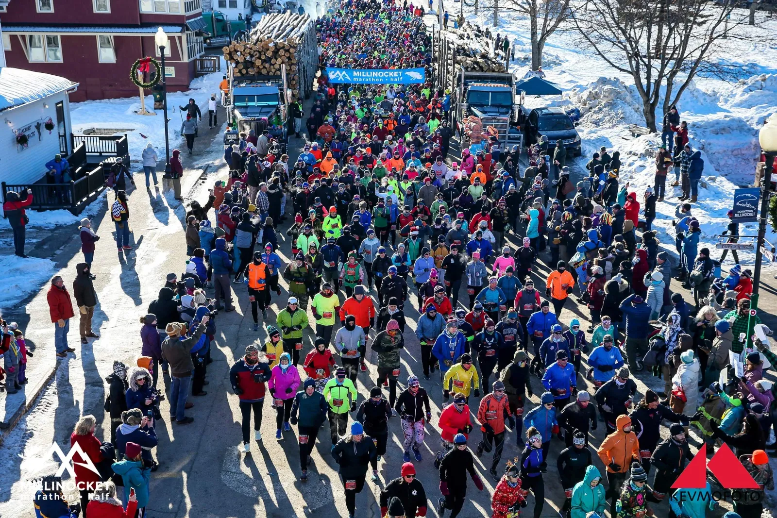 A large crowd of runners in colorful winter gear begins a race on a snow-lined street, with large logging trucks visible in the background and a banner overhead reading 'MILLINOCKET Marathon Half'.