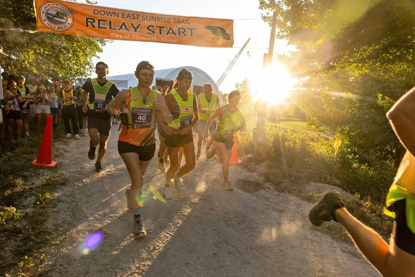 Runners starting a race under an orange banner that reads 'RELAY START' with the sun shining brightly in the background.