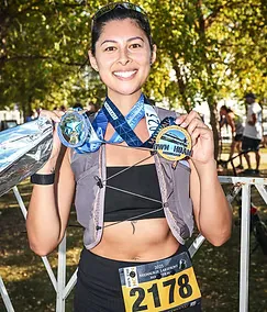 A smiling woman wearing two race medals around her neck and holding them up.