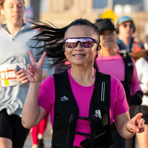 A female runner in hot pink gear and sunglasses smiles and gives a peace sign while running, with other runners in the background.