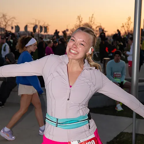 A smiling teenage girl in running attire with her arms outstretched, celebrating at a marathon event with other runners in the background.