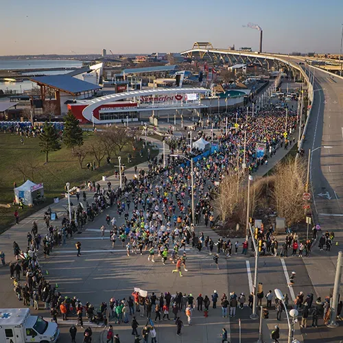 Aerial view of a large crowd of runners starting a marathon in an urban setting, with buildings and a stadium in the background.