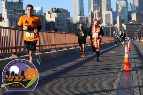 Runners in Halloween costumes, including a pumpkin and bacon, race across a bridge with a city skyline in the background.