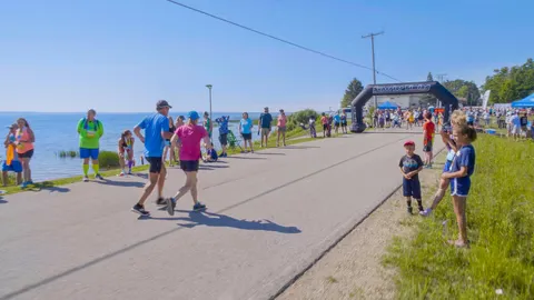Runners gather at the start line of a race with a large inflatable arch, with a body of water visible to the left and spectators on the right.