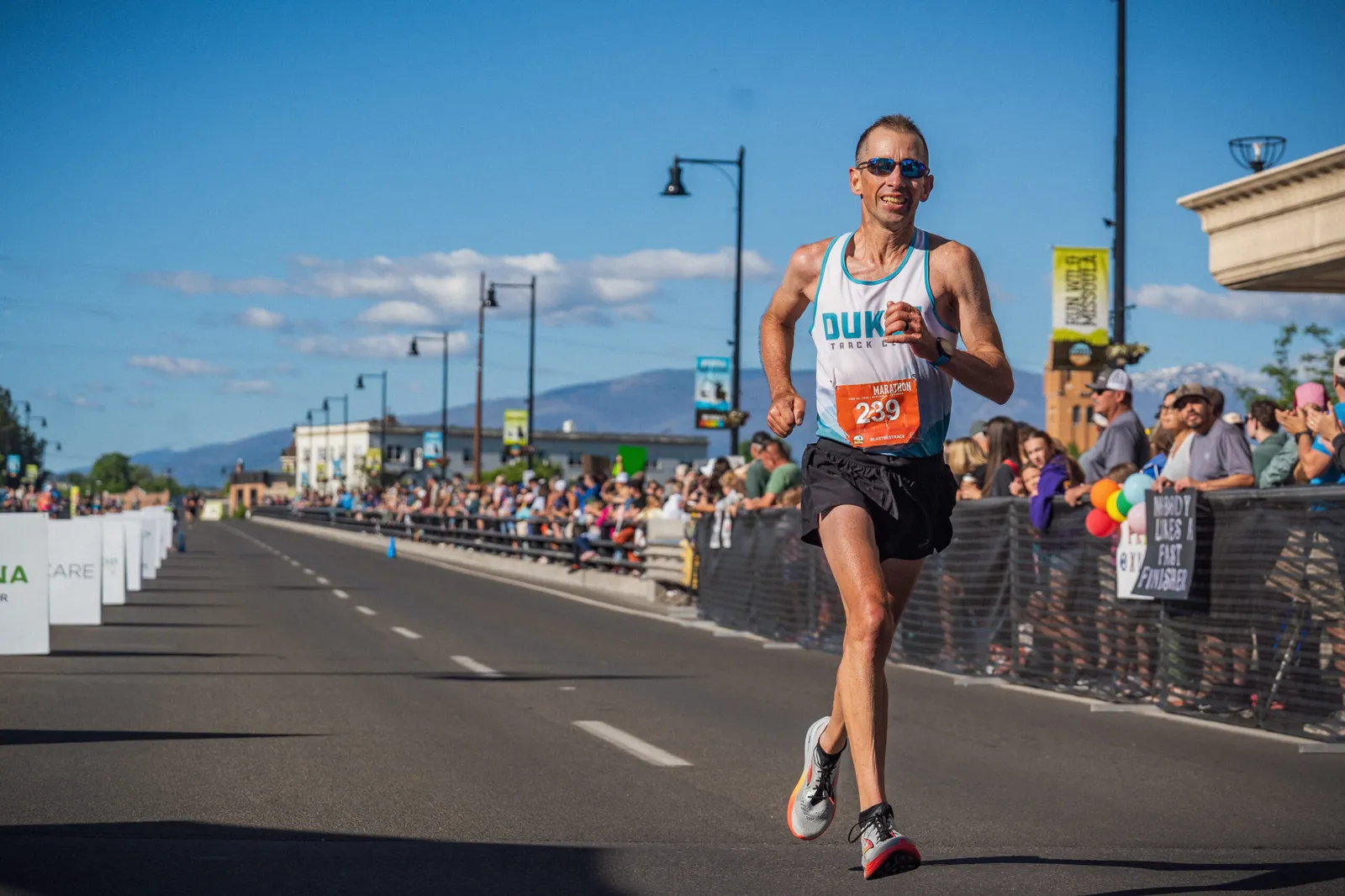 A male runner in a white tank top with 'DUKE TRACK CLUB' and a bib number '239' runs on a road with spectators cheering from the sidelines.