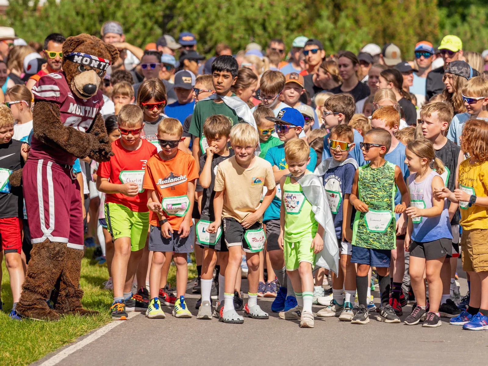 A group of children in running attire, some wearing capes, stand at the start line of a race, with a mascot in a bear costume interacting with them.
