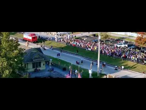 A large crowd of spectators cheers on runners at a race, with a train and a small building visible in the background.