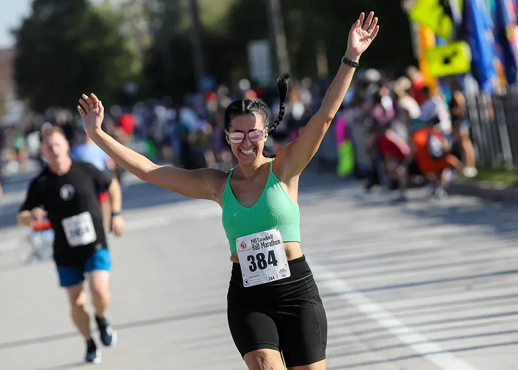 A female runner with her arms raised in celebration, wearing a green top and black shorts, with a race bib number 384.