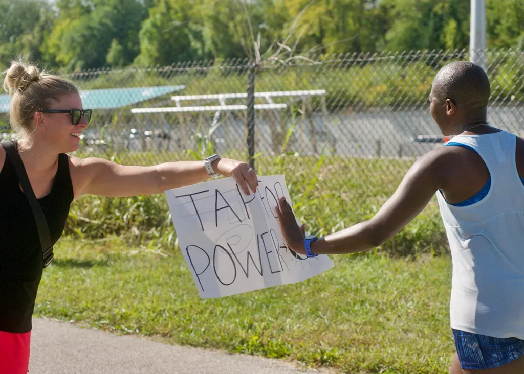 A runner holds out a sign that says 'TAP FOR POWER' to another runner during a race.