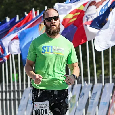 A male runner with a beard and sunglasses runs towards the camera, wearing a green t-shirt with race branding and a bib number 1000.
