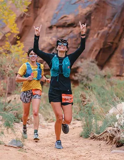 Two female runners on a desert trail, one in the foreground with arms raised in a rock-on gesture, both smiling.