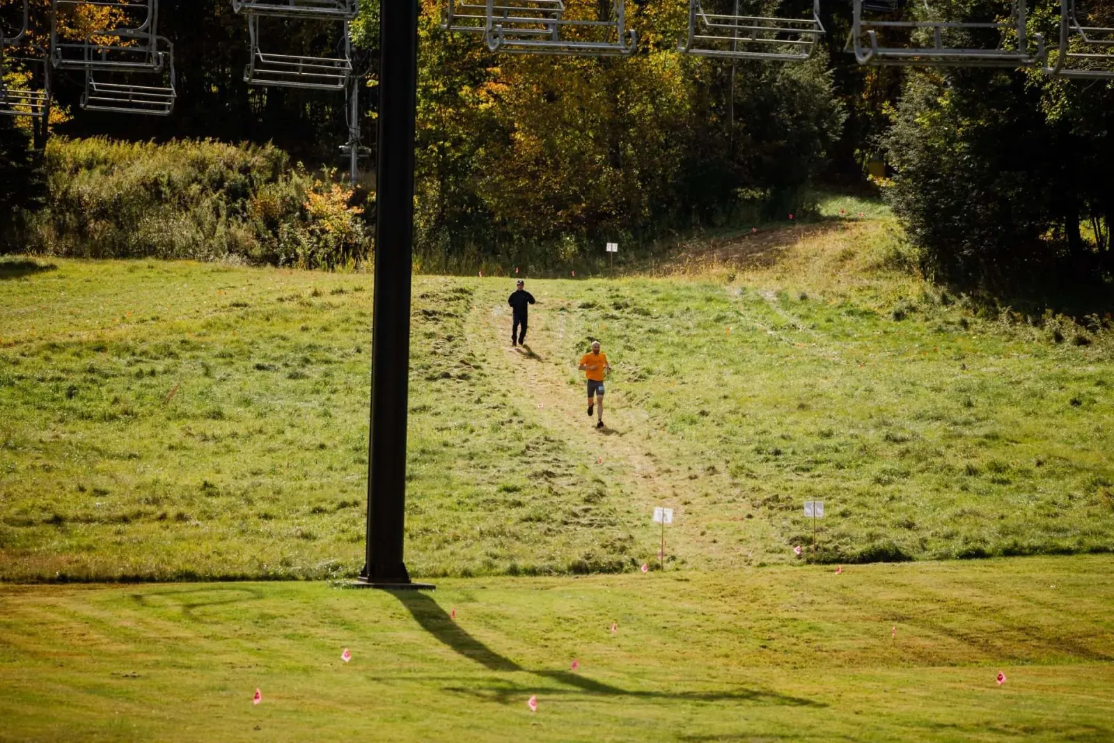 Two runners ascend a grassy hill on a trail, with ski lift chairs visible overhead and trees in the background.