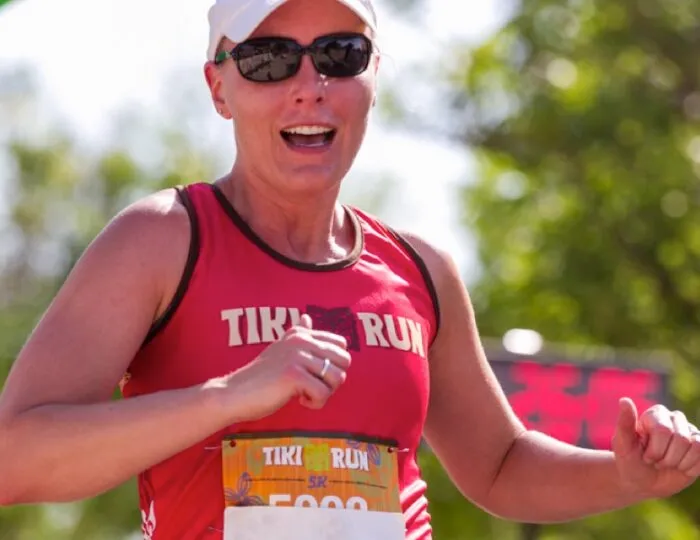A female runner wearing a red Tiki Run tank top and sunglasses smiles as she gives a thumbs up during a race.
