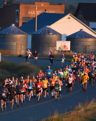 A large group of runners race down a road with buildings and silos in the background.