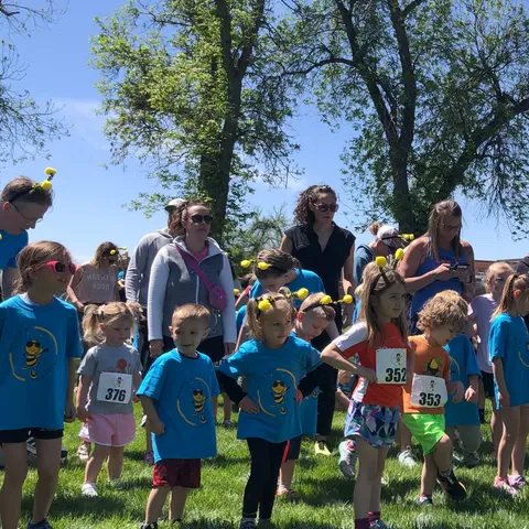 A group of children in blue shirts and bee headbands stand on a grassy field, some with race bibs, appearing ready for a race.