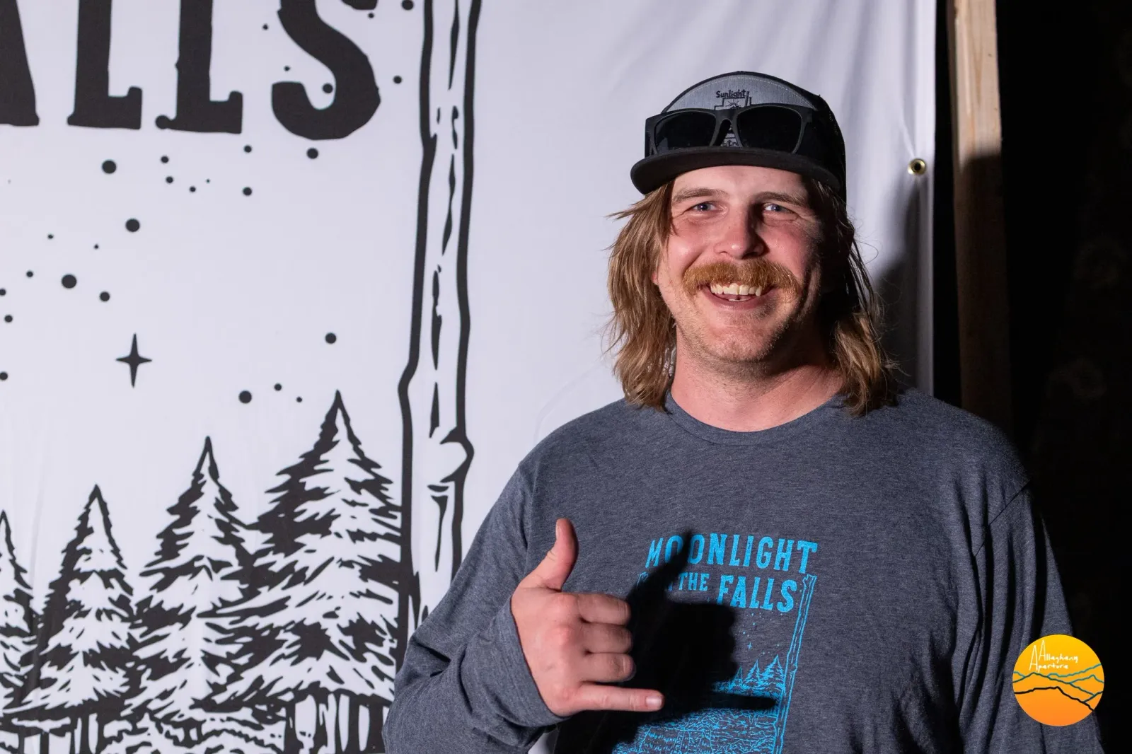 A smiling man with a mustache and a baseball cap gives a shaka sign while wearing a long-sleeved shirt with 'Moonlight on the Falls' printed on it.