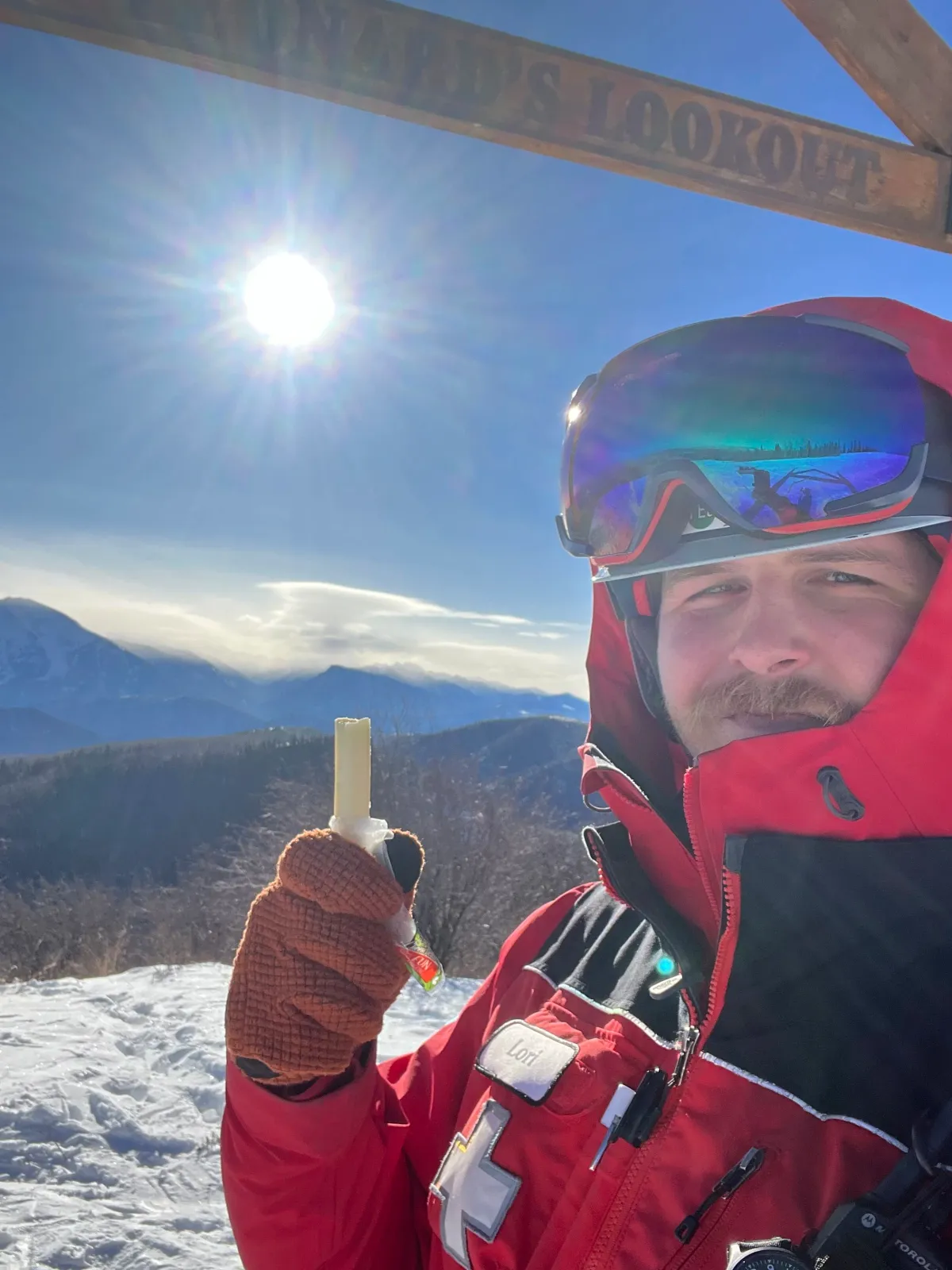 Selfie of a person in a red jacket and ski goggles, holding a small tube, with a scenic mountain landscape and a sign that reads 'MONARD'S LOOKOUT' in the background.