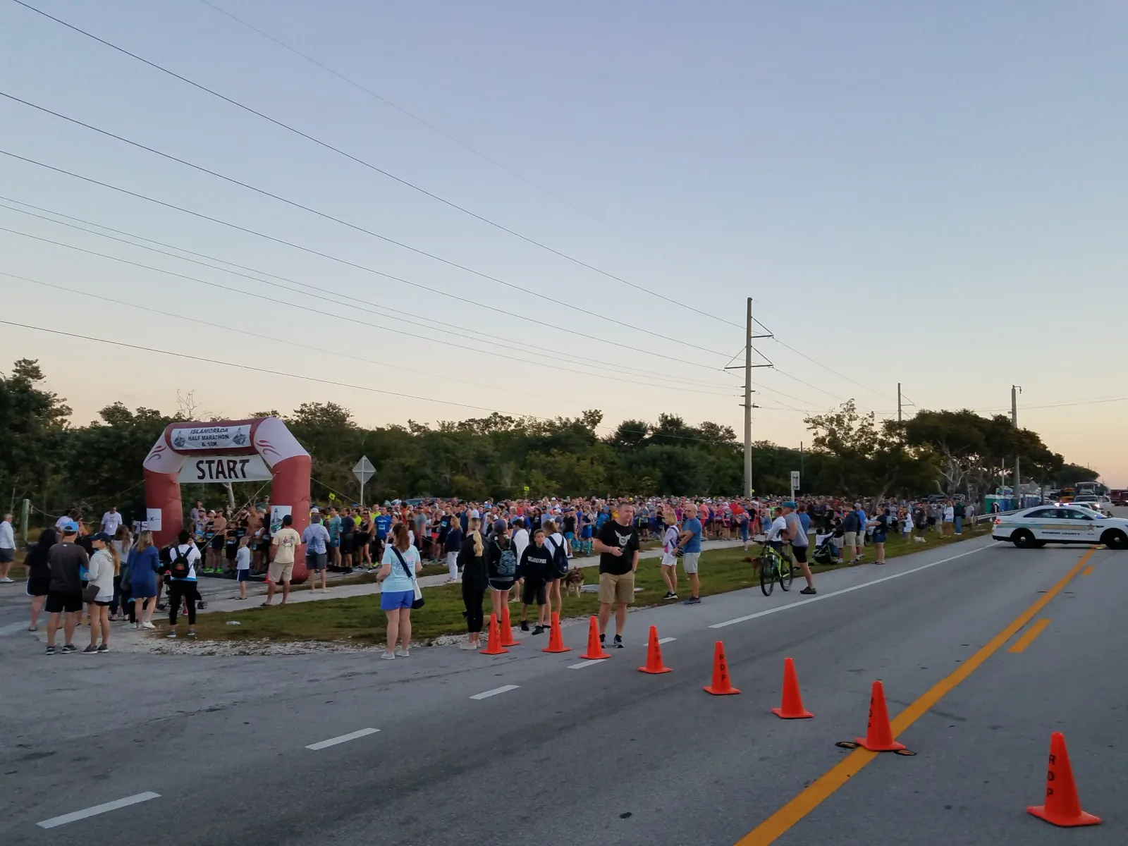 A large crowd of runners gathers at the start line under an inflatable archway, with cones marking the course and a police car present.