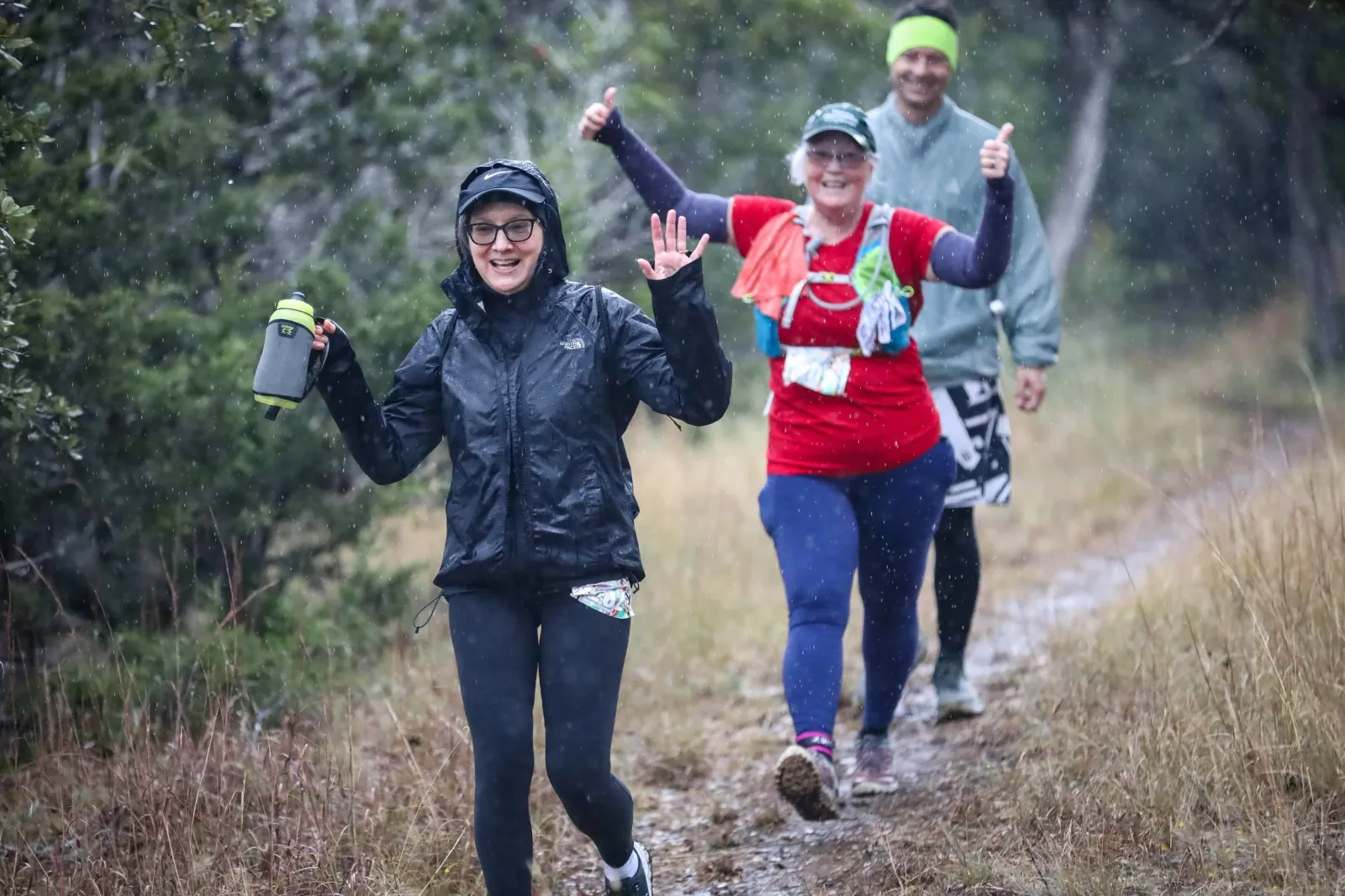 Three runners smiling and giving thumbs up while running on a trail in the rain.