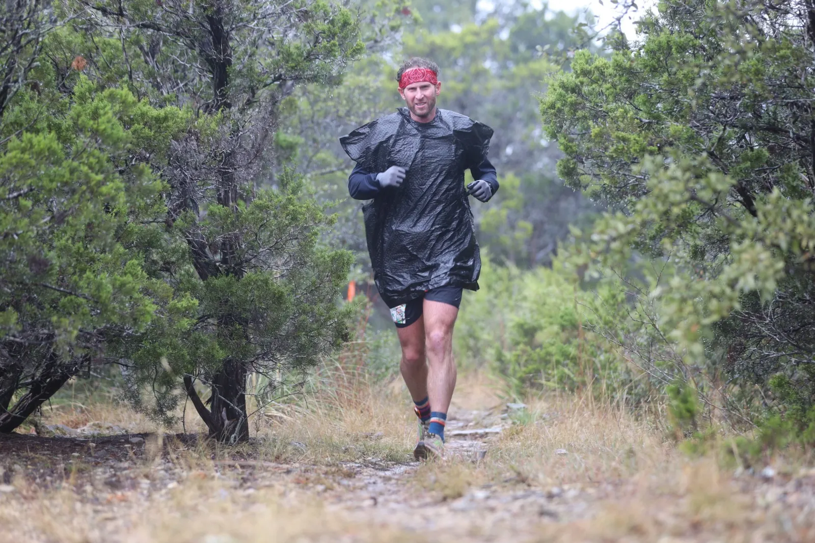 A male runner wearing a red bandana and a black poncho runs on a trail in a wooded area.