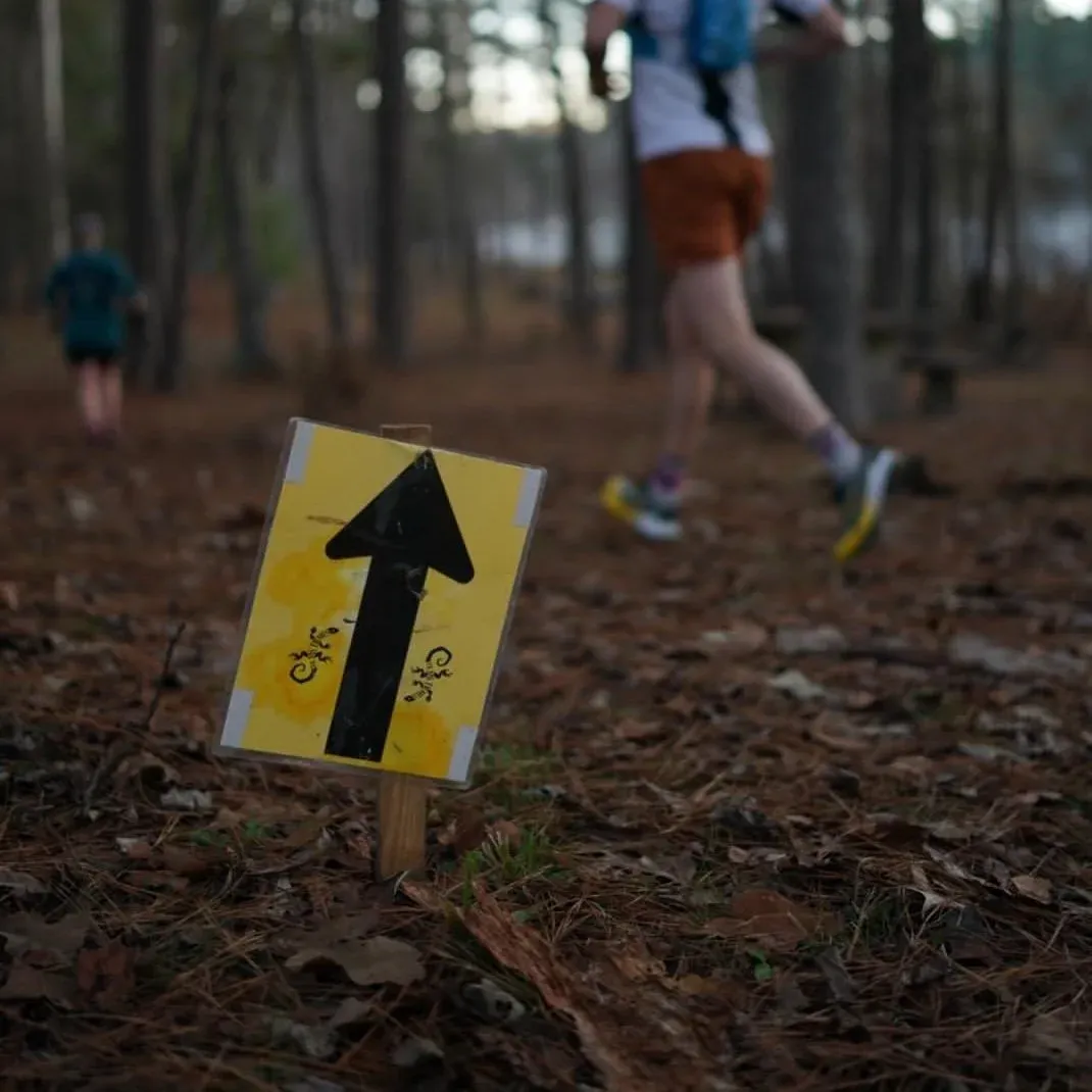 A runner on a wooded trail passes a yellow directional sign with a black arrow.