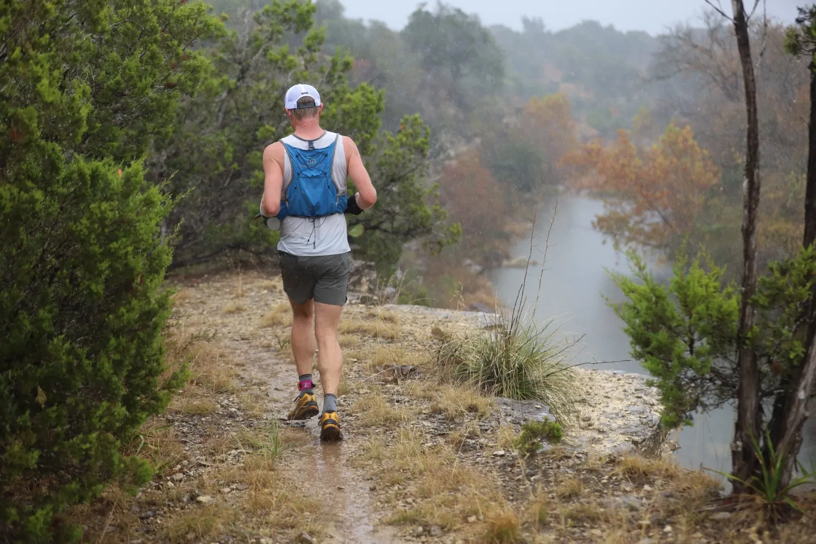 A runner with a blue hydration pack runs on a wet trail with trees and a body of water in the background.