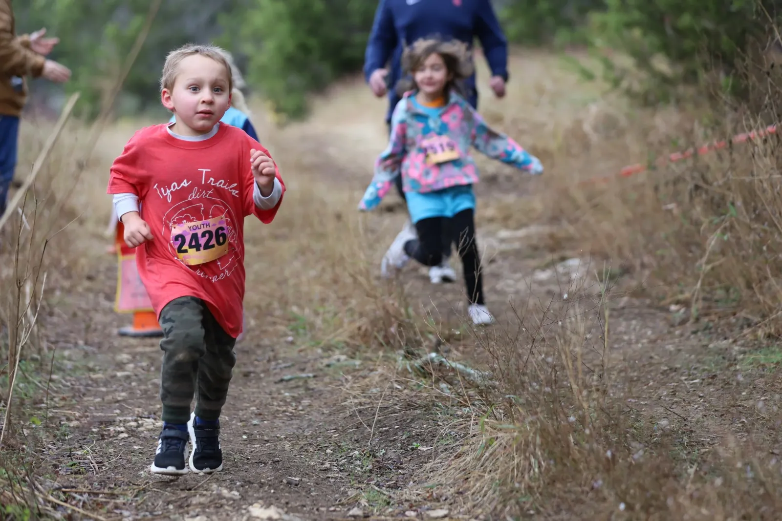 A young boy in a red shirt with bib number 2426 runs on a dirt trail, with other runners and spectators in the background.